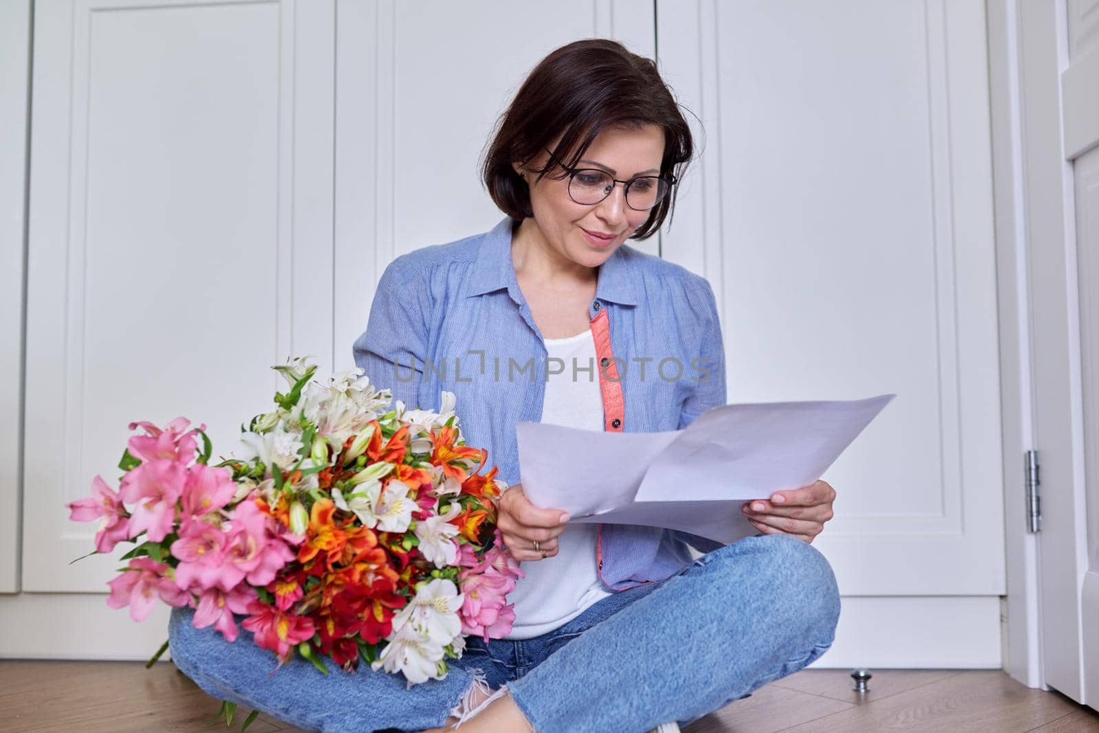 Middle-aged smiling female with a bouquet of flowers reading paper. Holiday, date, anniversary, good business news, positive letter, congratulations concept