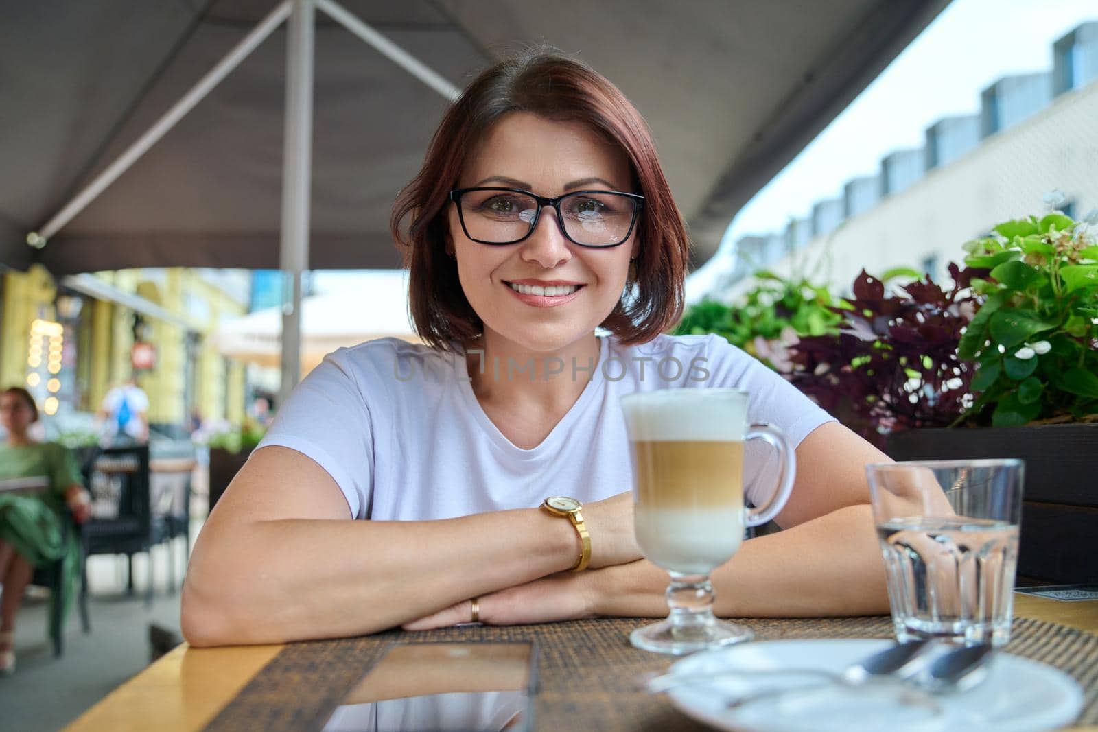 Portrait of smiling middle aged woman looking at camera, 40s female wearing glasses sitting in an outdoor cafe with cup of coffee. Lifestyle, urban style, middle-aged people concept