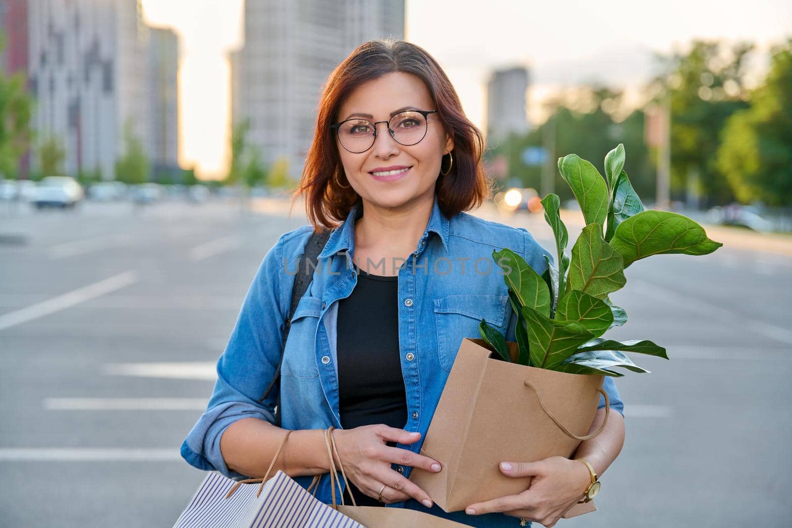 Middle aged woman with paper shopping bags with buying plant, looking at camera by VH-studio