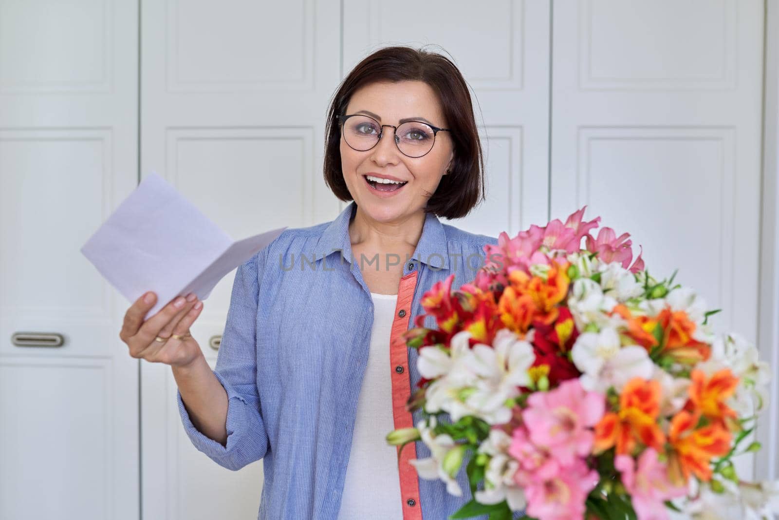 Happy middle age woman with bouquet of flowers looking in camera with paper text congratulation by VH-studio