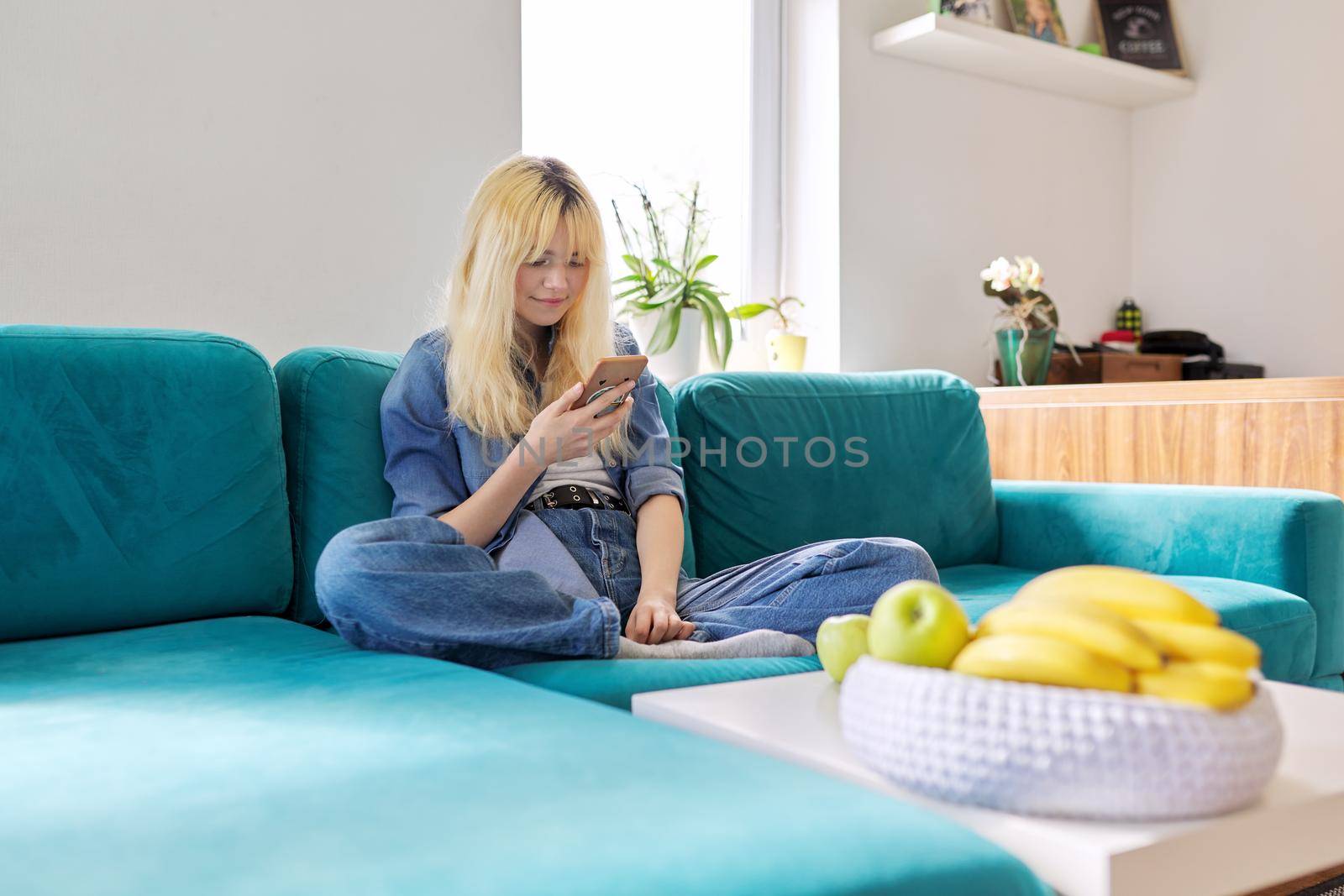 Smiling happy female teenager 16 years old sitting at home on couch looking at smartphone screen, texting, browsing blog, social networks, chatting with friends online