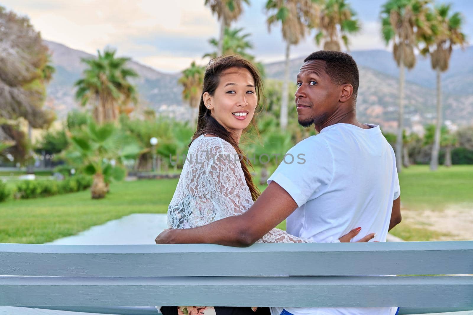 Beautiful young multi-ethnic couple sitting together on bench in tropical park. Loving asian woman and african man embracing. Love relationship, happiness, multicultural family, tourism travel, people
