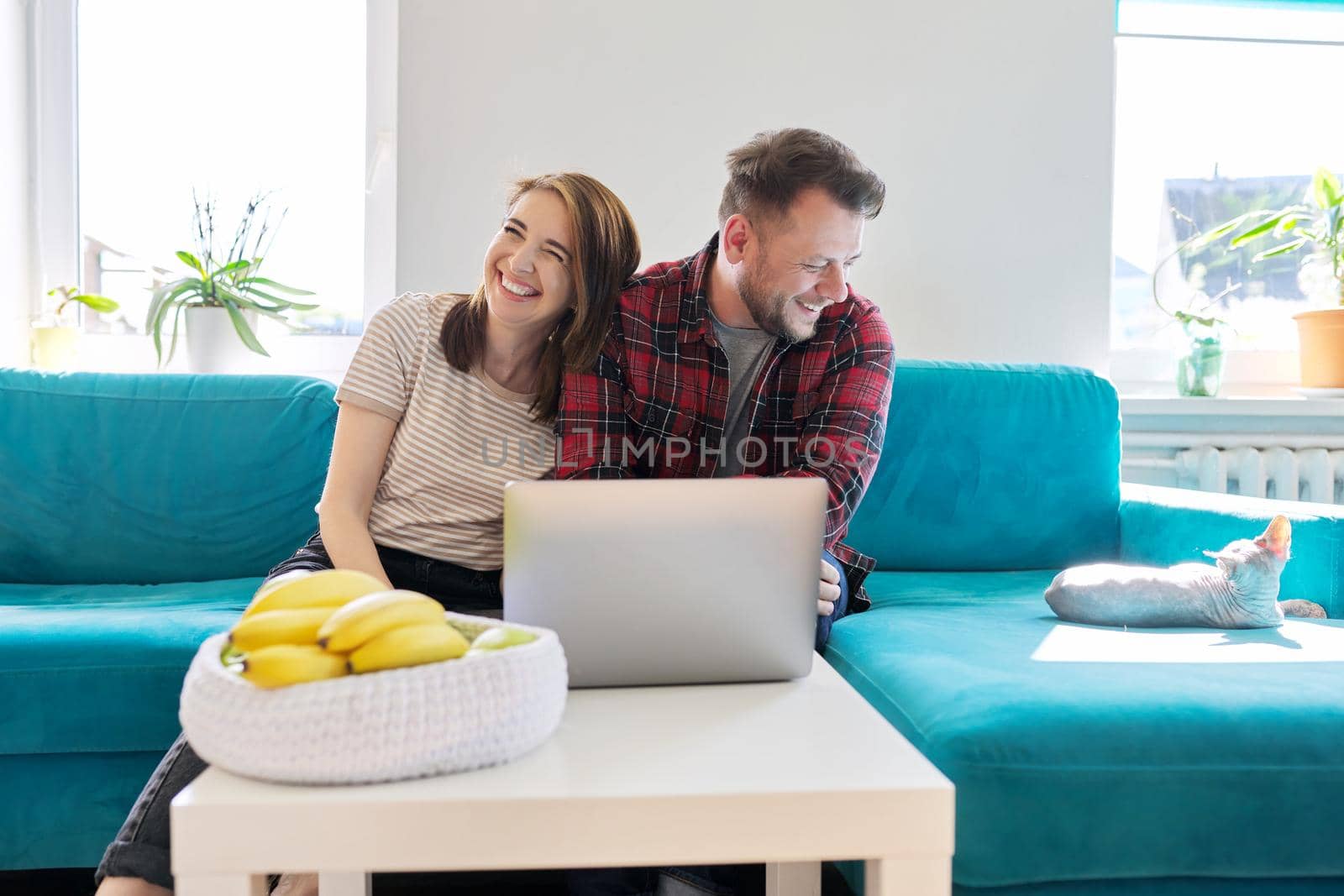 Smiling laughing positive couple 40 years old, husband and wife looking at laptop monitor sitting together at home on sofa in living room, real people life