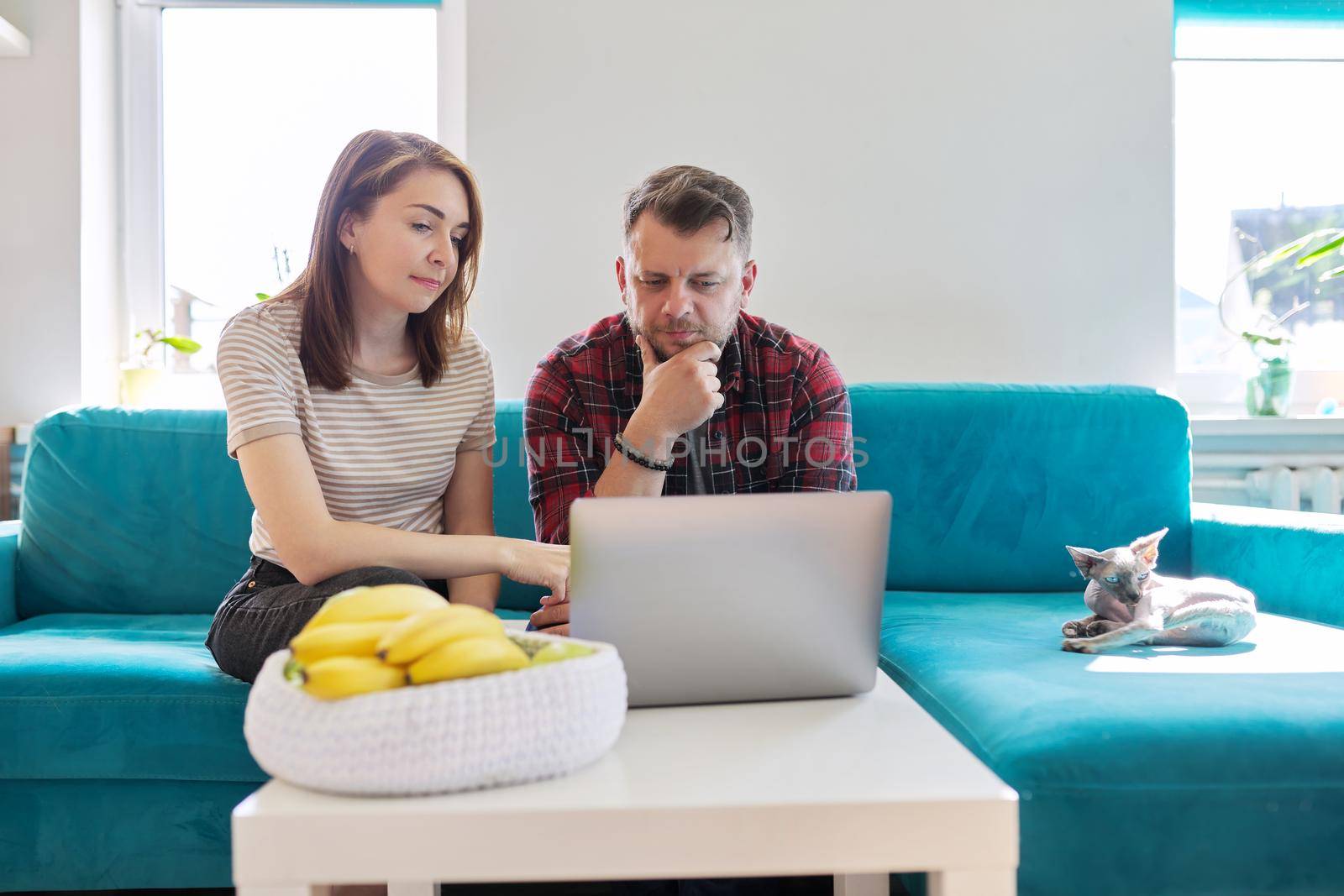 Serious middle-aged couple husband and wife with laptop sitting at home on couch. Man and woman attentively listening and looking at computer monitor, online meeting, video consultation, chat conference