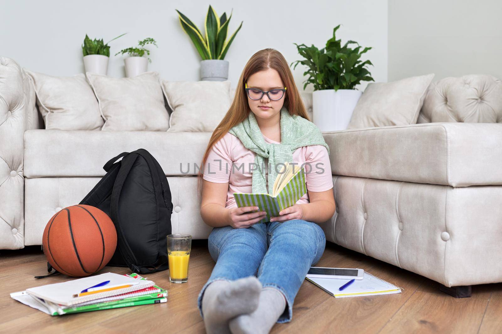 Lifestyle, education children 12, 13 years old concept. Teenage girl sitting at home on the living room floor reading book with backpack, books, school notebooks, basketball