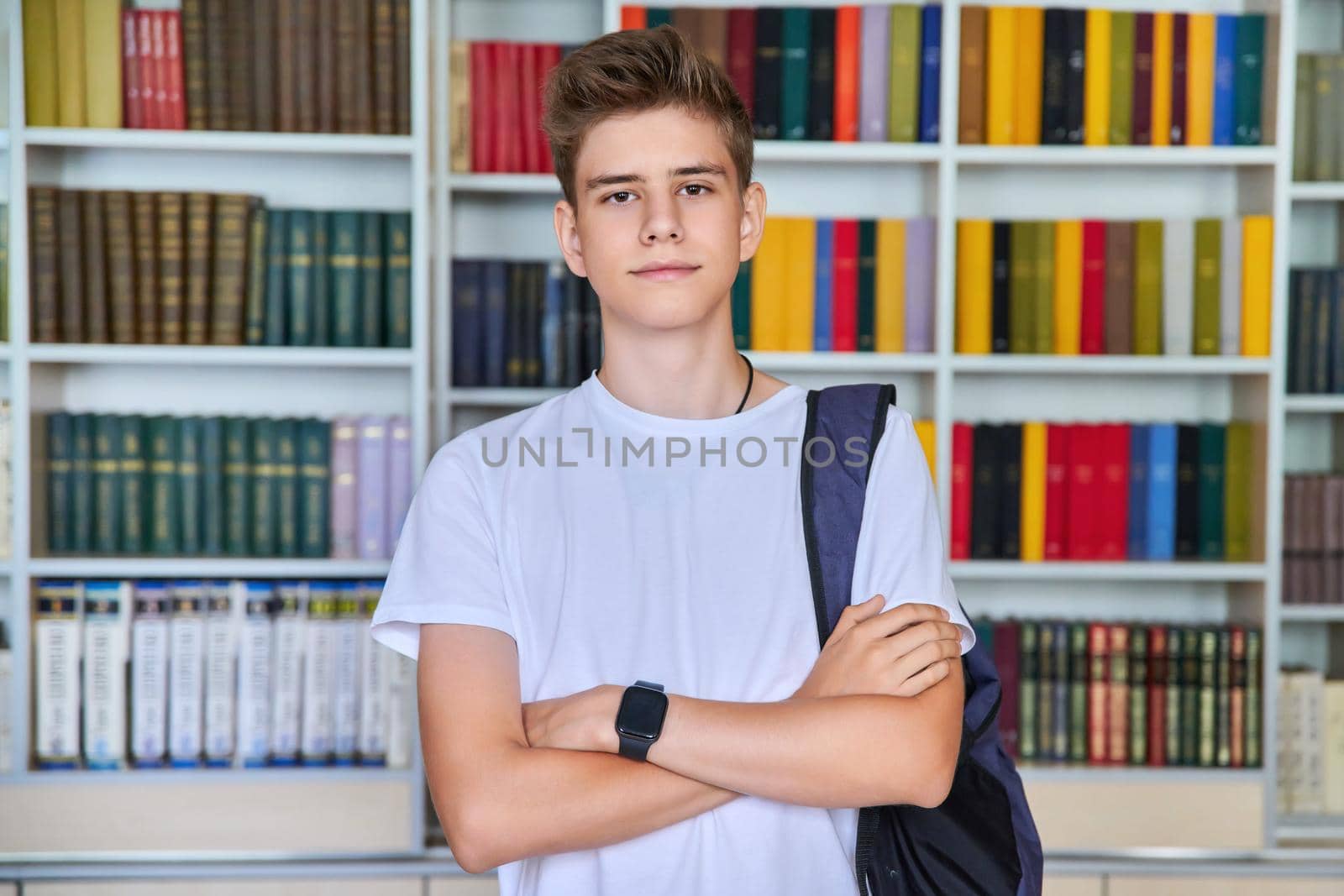 Single portrait of serious confident male student teenager with backpack with crossed arms looking at camera in the library. School, education, knowledge, adolescence concept