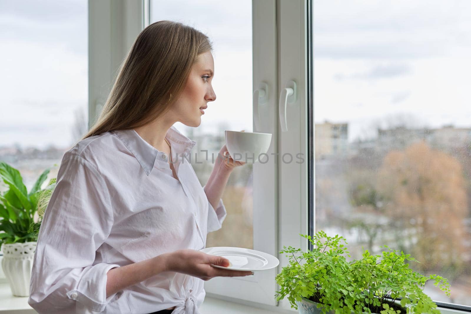 Portrait of young beautiful woman in white shirt with cup of coffee near window. Blonde female relaxing looking out the window of winter autumn city
