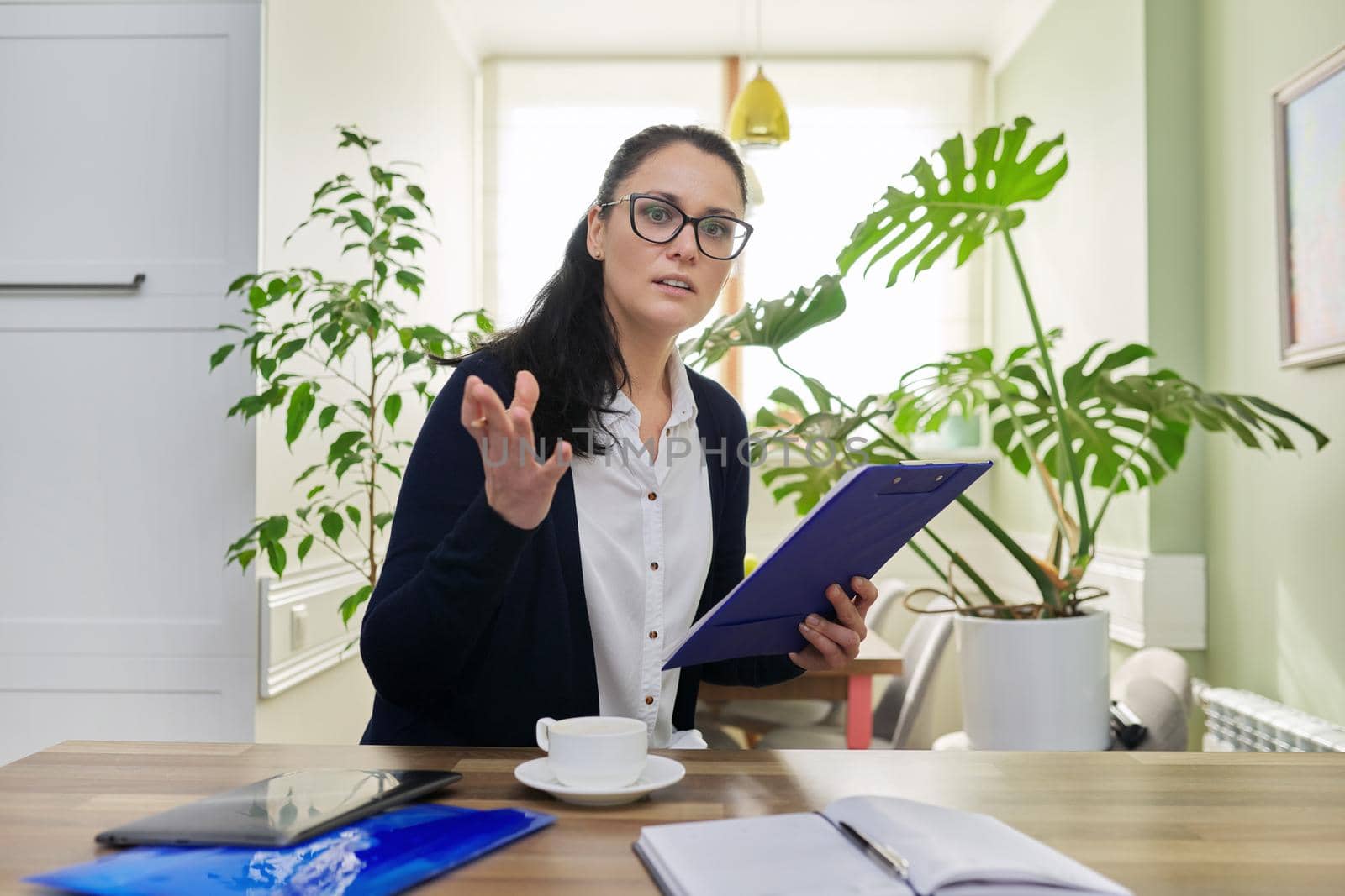 Business woman looking at camera talking, holding papers documents in hands, home interior background. Online consultation, remote work, paperwork, internet, technologies in business, education