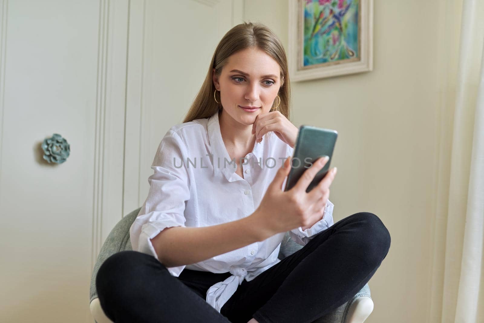 Emotional laughing young beautiful woman looking at smartphone screen, relaxing sitting at home in chair. Video call, emotional communication, joy, happiness, surprise, technology in everyday life