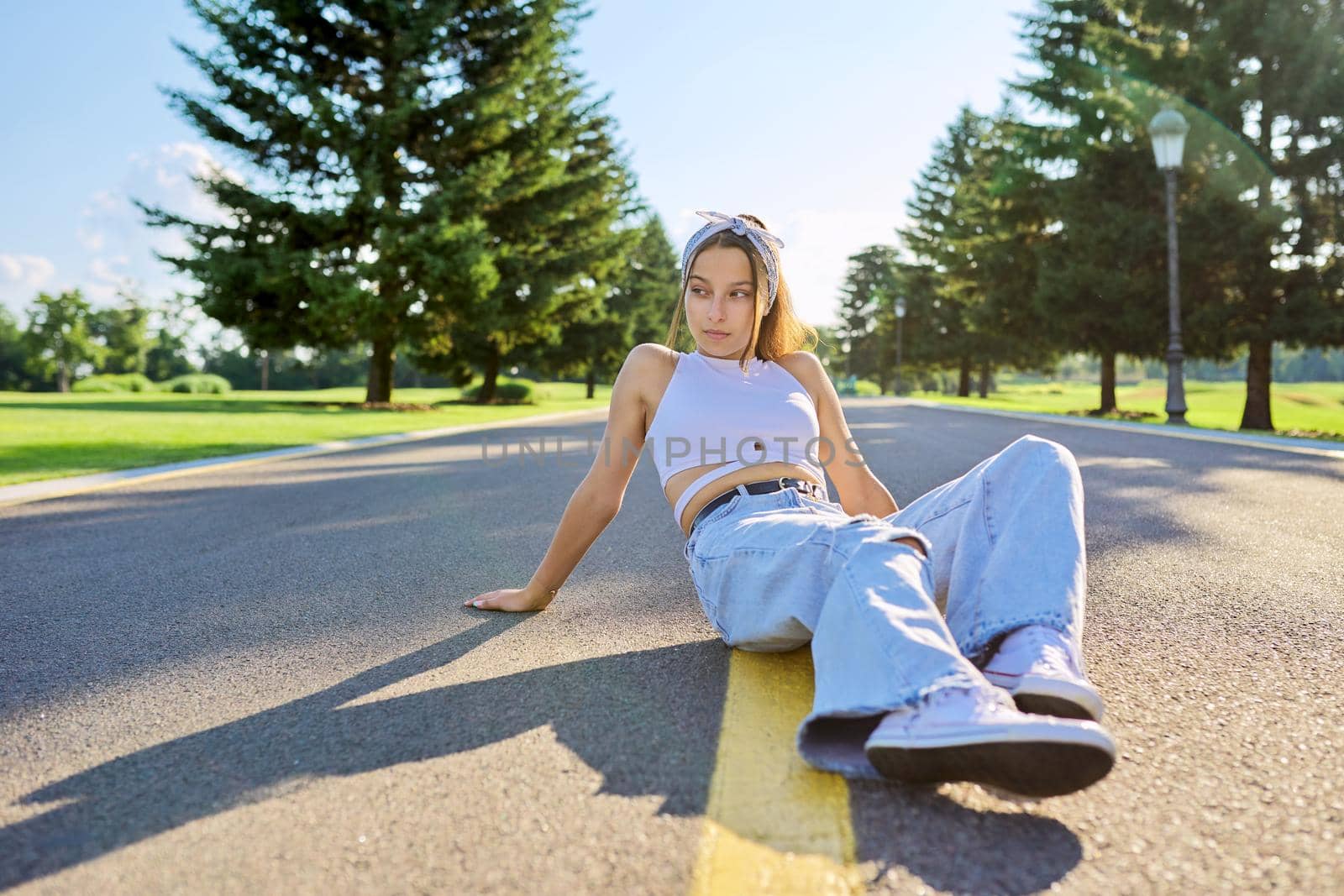 Fashionable trendy teenage hipster female sitting on road in park on sunny summer day. Young stylish model, outdoor. Youth, teens, summer, fashion, street style, lifestyle concept