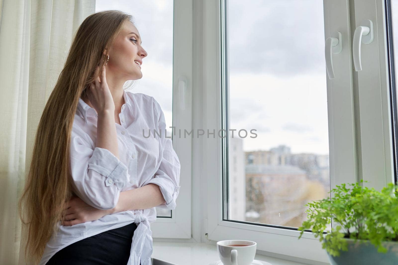 Portrait of young beautiful woman in white shirt with cup of coffee near window. Blonde female relaxing looking out the window of winter autumn city