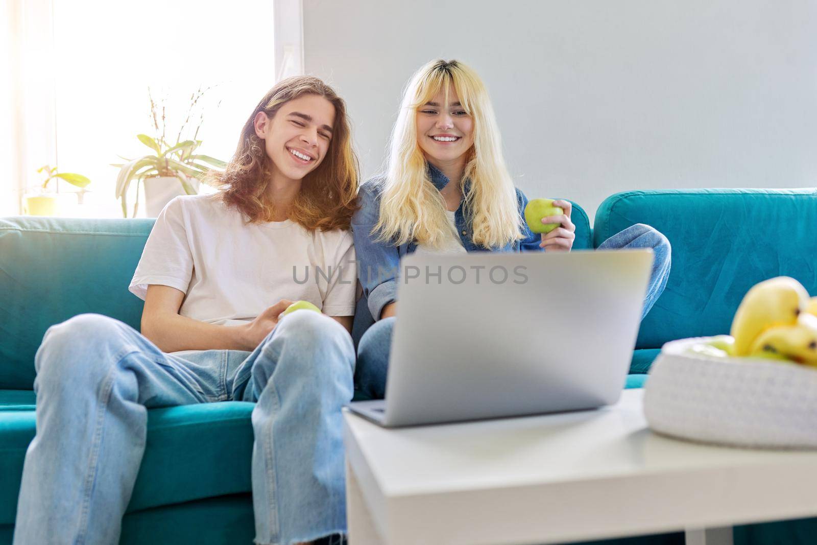 Laughing couple of teenagers having fun, sitting on couch, looking at laptop by VH-studio