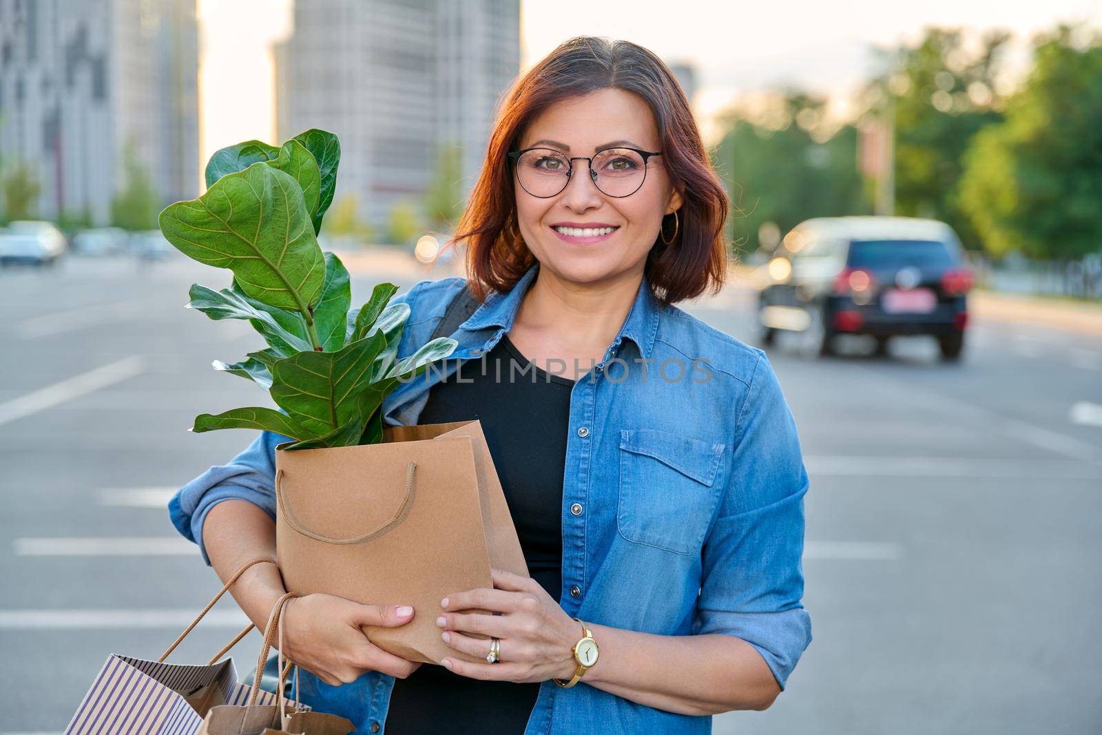 Middle aged woman with paper shopping bags with buying plant, looking at camera by VH-studio