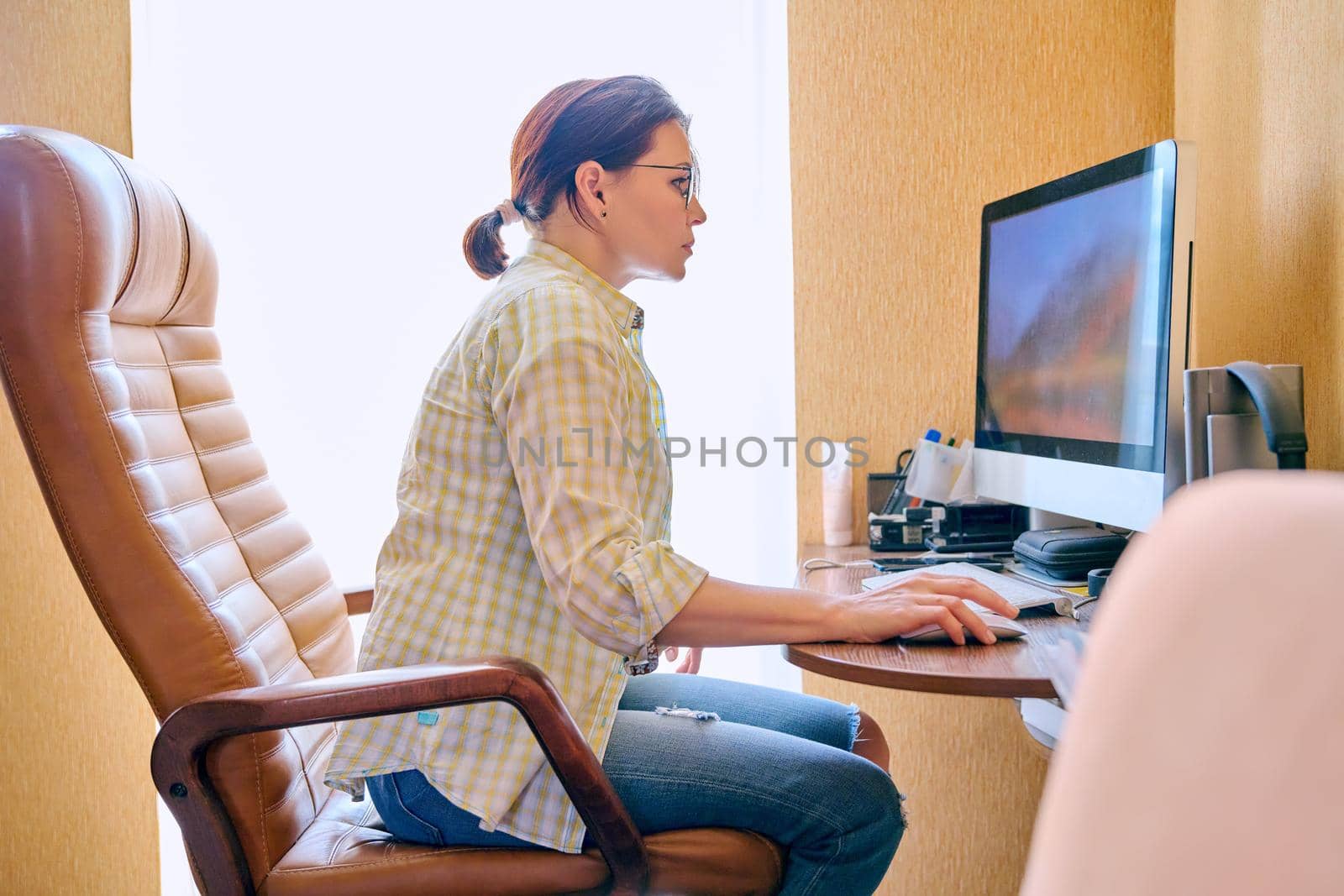 Middle-aged woman freelancer at home workplace. Serious female wearing glasses in casual clothes working remotely using computer sitting on armchair near window