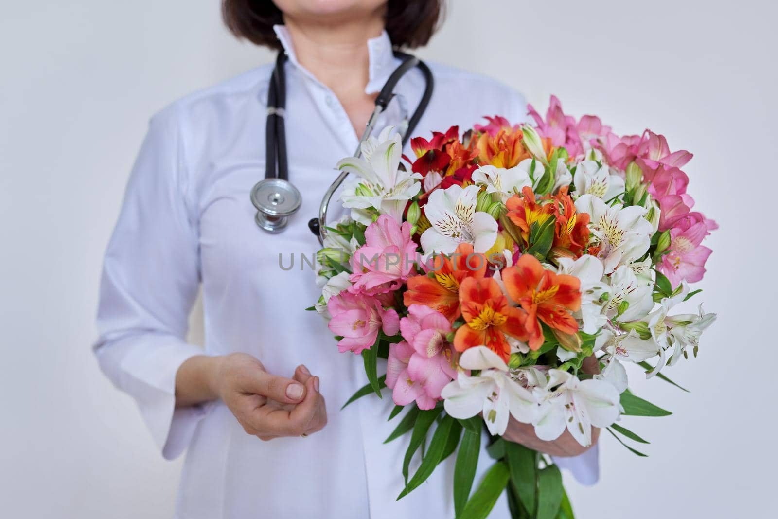 Close-up of bouquet of flowers in hands of female doctor. Medic, pharmacist in white uniform with stethoscope on light background. International, National Doctor's Day, Nurse's Day, Health Day