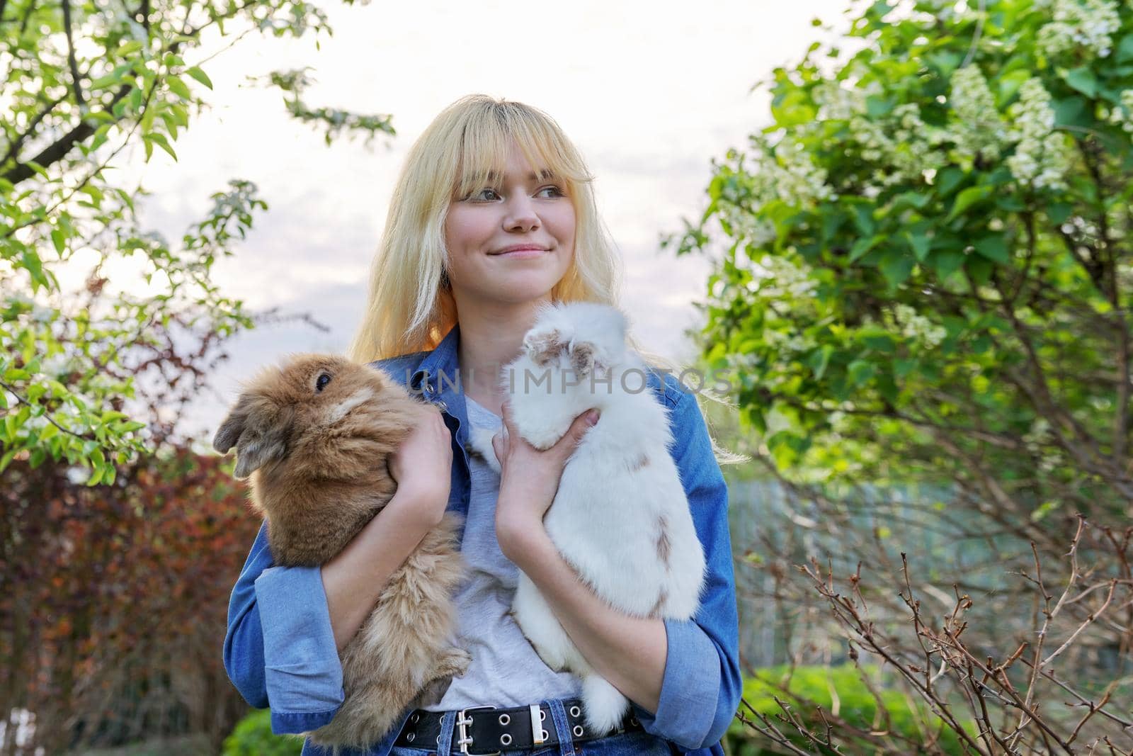 Beautiful smiling girl teenager with a couple of decorative rabbits in their hands. Blonde female in denim clothes with two bunnies in a spring blooming garden