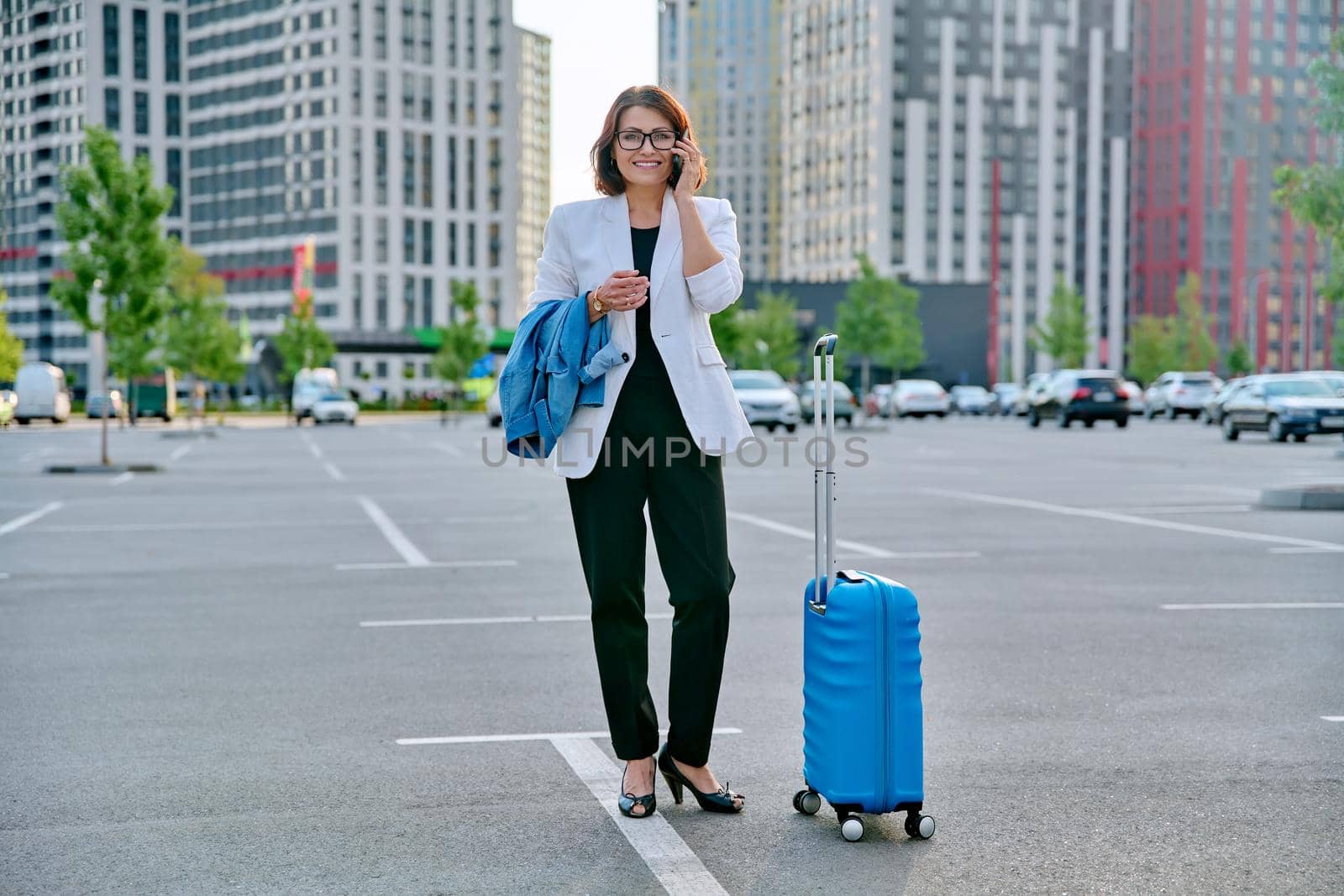 Middle aged businesswoman with suitcase talking on mobile phone in outdoor parking, urban style, city architecture background. Business travel trip, business people of mature age, success, modern city