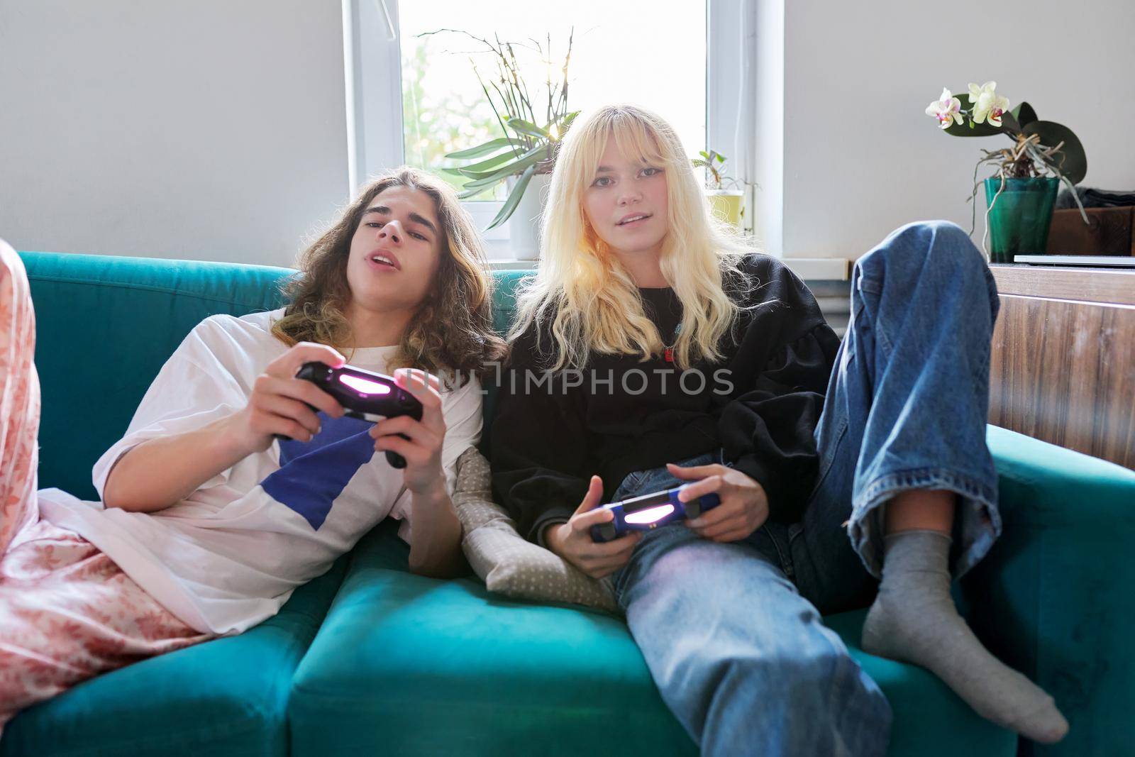 Couple of 15, 16 years old teenage friends playing with video game console, sitting at home on couch, having fun together. Real people, teenagers lifestyle, virtual computer games concept