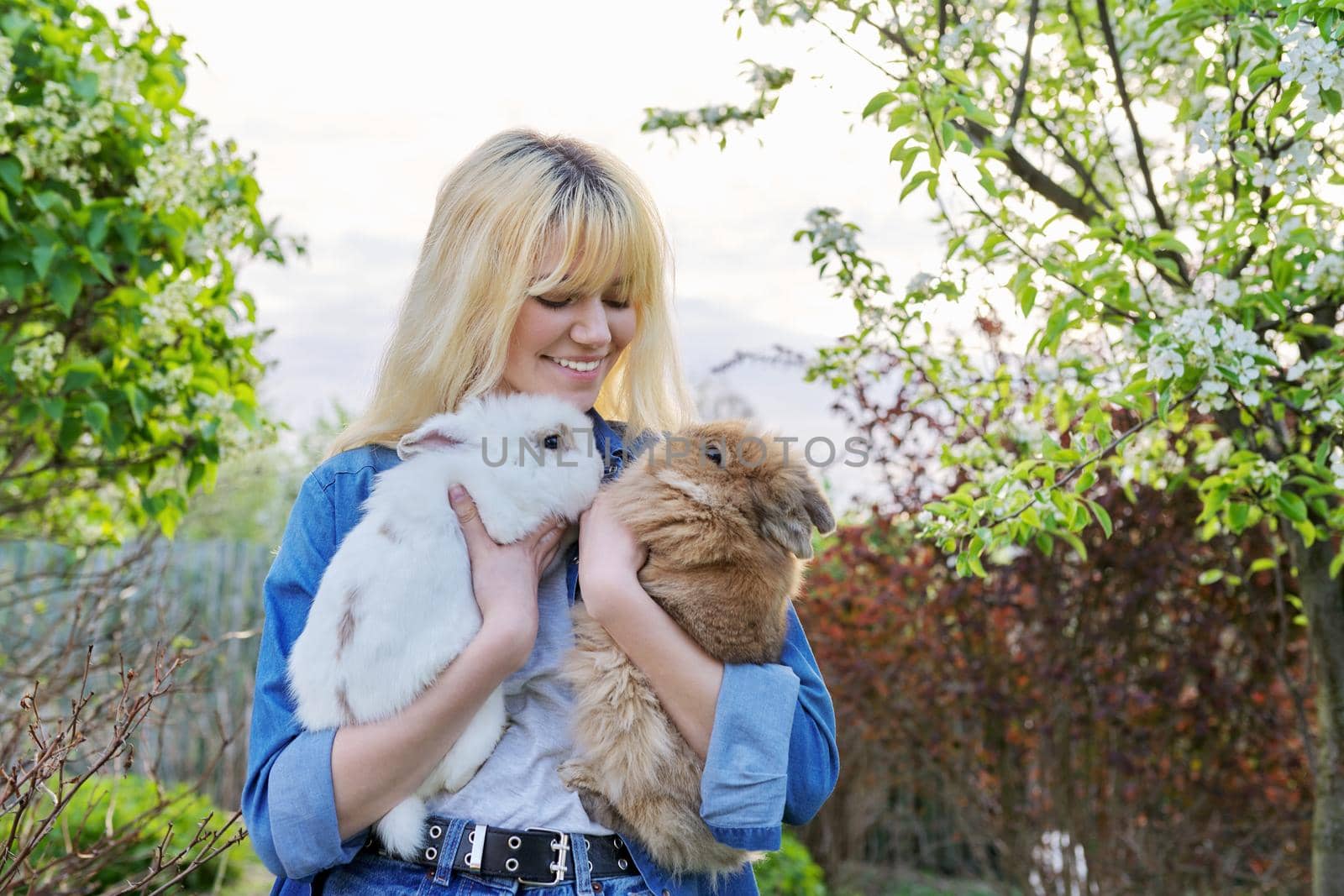 Beautiful smiling girl teenager with a couple of decorative rabbits in their hands. Blonde female in denim clothes with two bunnies in a spring blooming garden