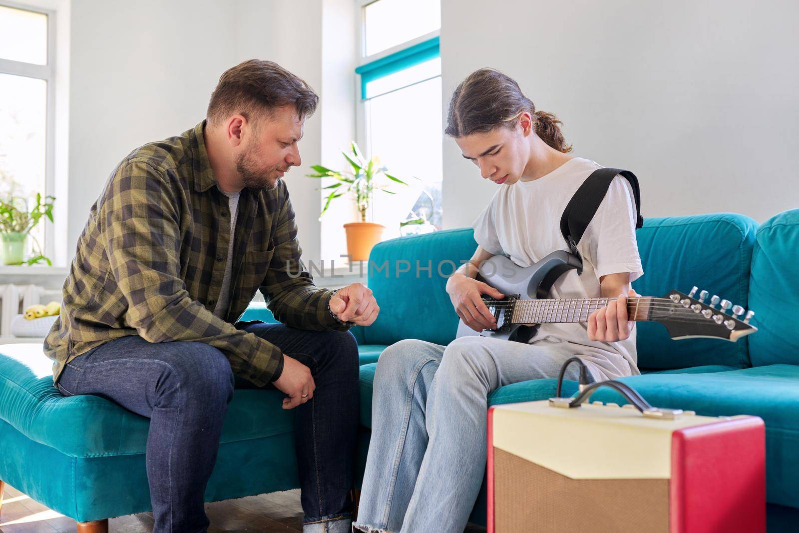 Father teaches his teenage son to play the electric guitar. Family sitting together at home in living room on couch. Parent-child relationship, lifestyle, creativity, music, teenagers concept