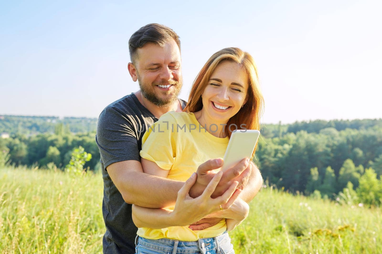 Beautiful happy adult couple taking selfie on smartphone, sunny summer day meadow forest sky background. Relationships, happiness, relaxation, love, people 30s 40s age