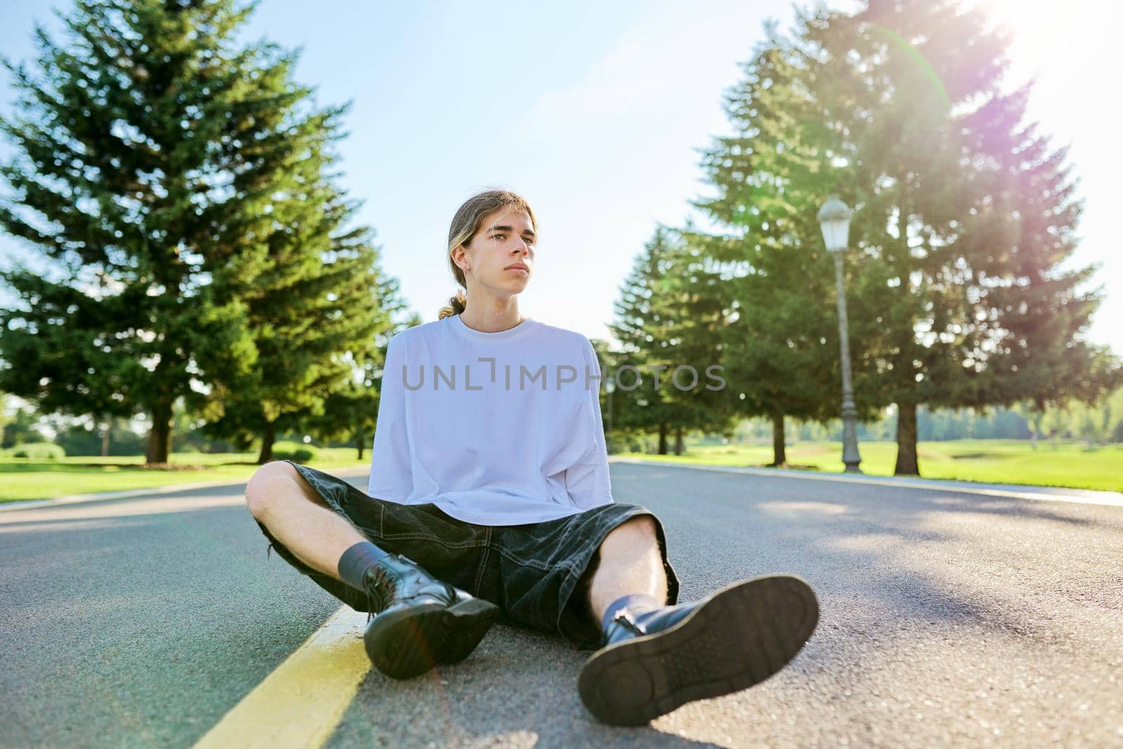 Fashion portrait of hipster teenager guy sitting on road. Serious posing young male with long hair wearing white t-shirt shorts boots on sunny summer day. Youth, adolescence, people, lifestyle concept