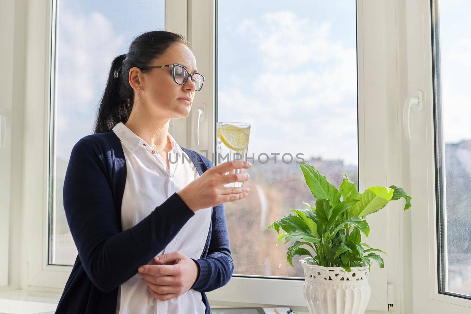 Business woman with glass of water with lemon. Middle-aged female in glasses, white shirt cardigan near window, resting enjoying healthy vitamin water