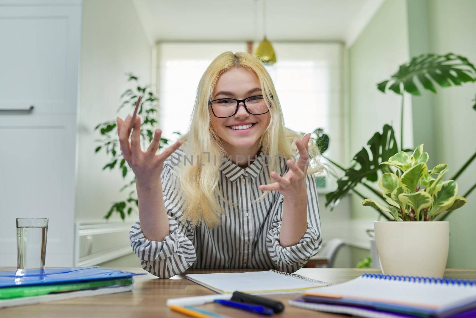 Portrait of teenager female student looking at camera, talking smiling, sitting at home at the table with notebooks textbooks. Looks in webcam, online education, video lesson, conference, e-learning