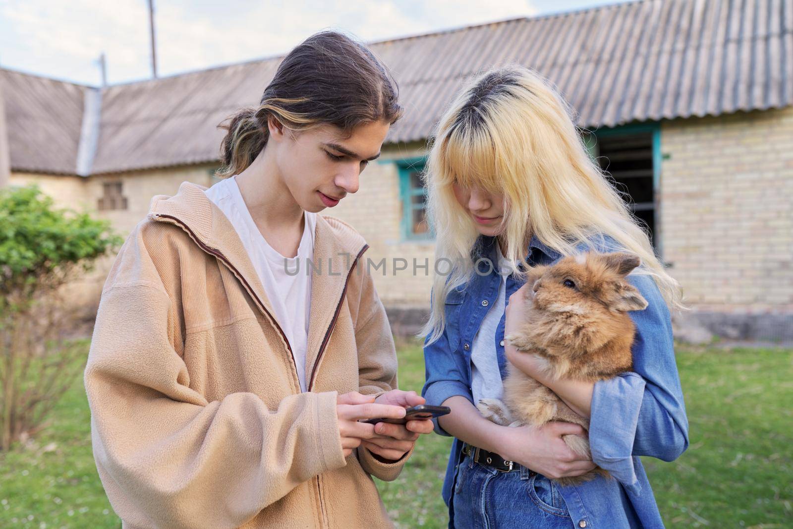 Teenagers guy and girl with decorative rabbit in their hands talking and looking at smartphone screen, on rural farm, spring blooming garden background, countryside, domestic animals