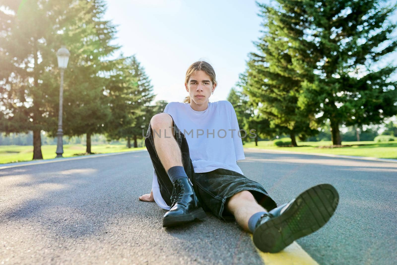 Fashion portrait of hipster teenager guy sitting on road. Serious posing young male with long hair looking in camera,in white t-shirt shorts boots on summer day. Youth, adolescence, people, lifestyle