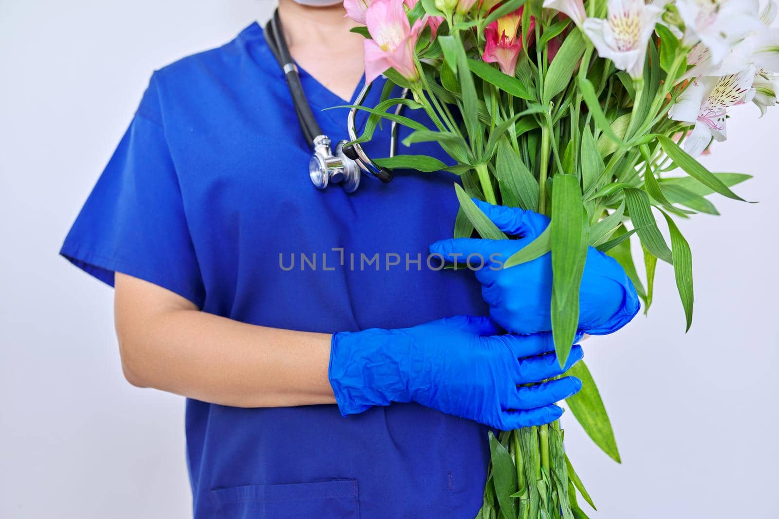 Health day, close-up bouquet of flowers in the hands of doctor nurse. Female medic in blue medical uniform, protective gloves with stethoscope on light background