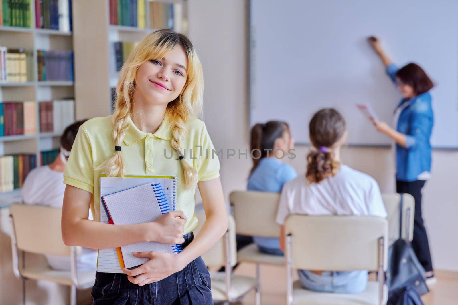 Female teenager student smiling looking at camera, group of pupils and teacher in library. High school, college, knowledge, education, adolescence concept