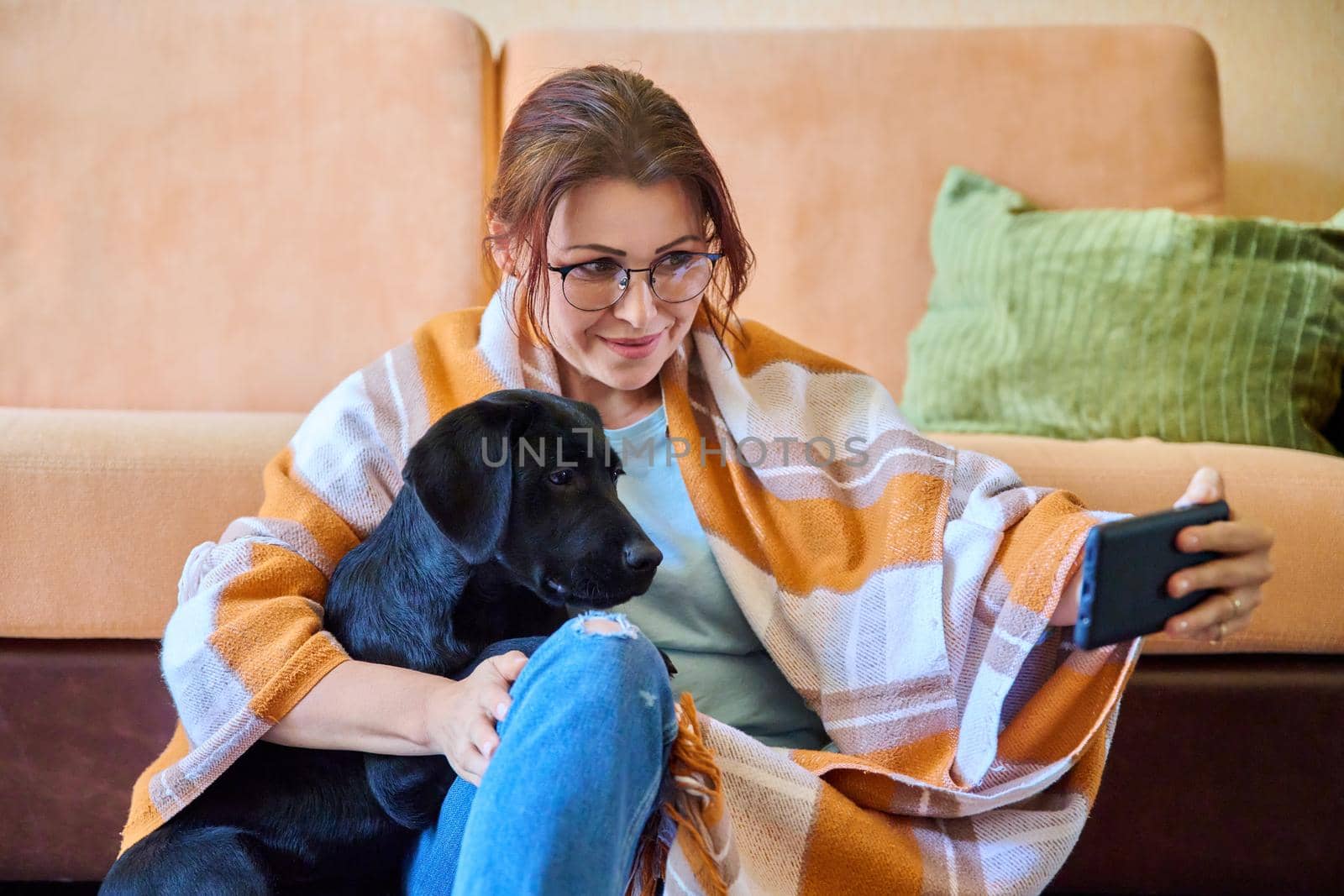 Winter autumn portrait of smiling woman and dog at home. Middle-aged female in warm blanket hugging her pet black labrador puppy takes selfie on smartphone. Lifestyle, love, pets, 40s people concept