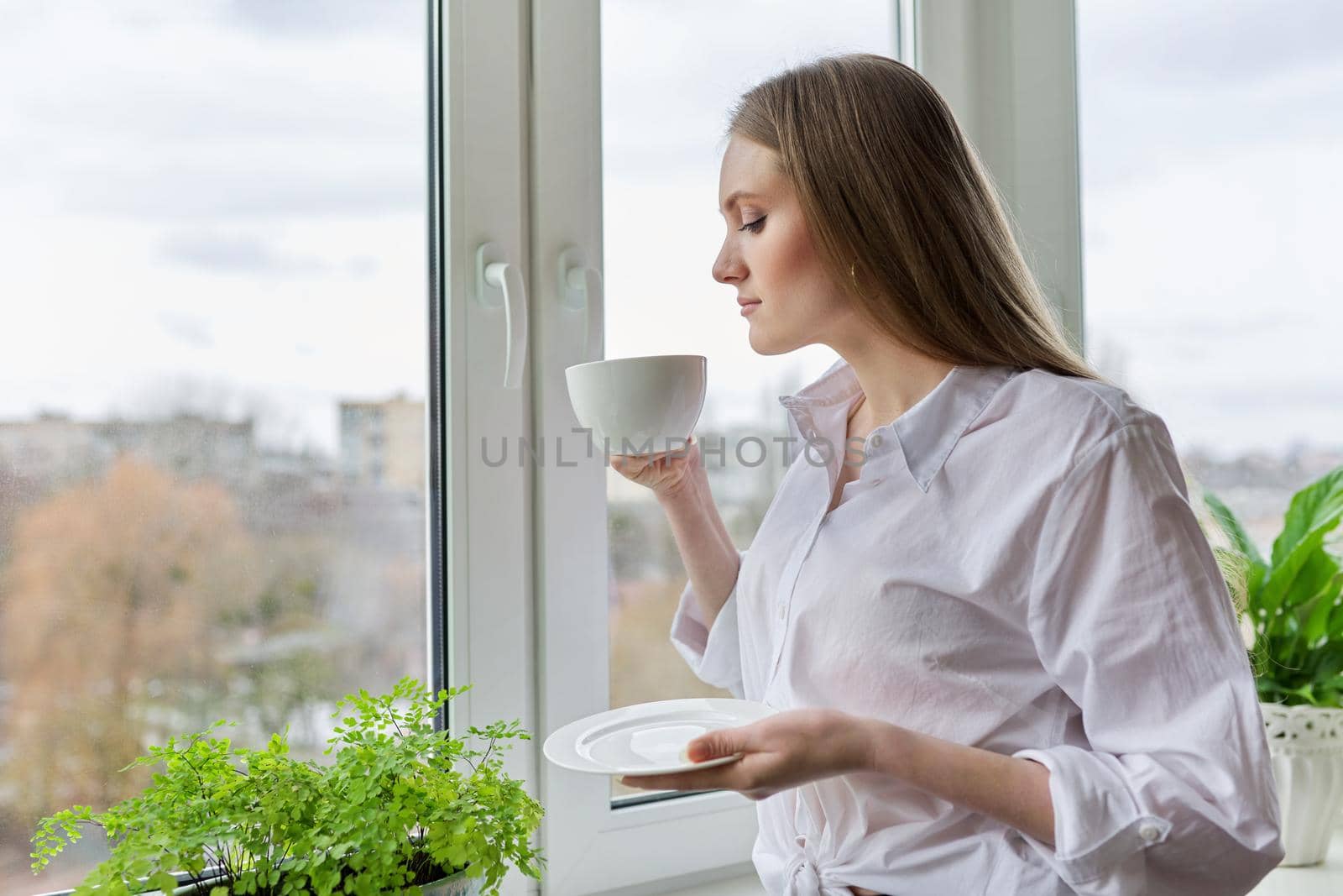 Portrait of young beautiful woman in white shirt with cup of coffee near window. Blonde female relaxing looking out the window of winter autumn city