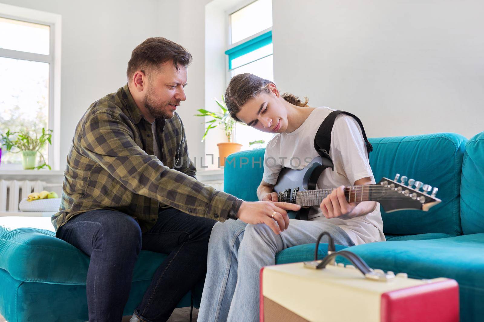 Father teaches his teenage son to play the electric guitar. Family sitting together at home in living room on couch. Parent-child relationship, lifestyle, creativity, music, teenagers concept