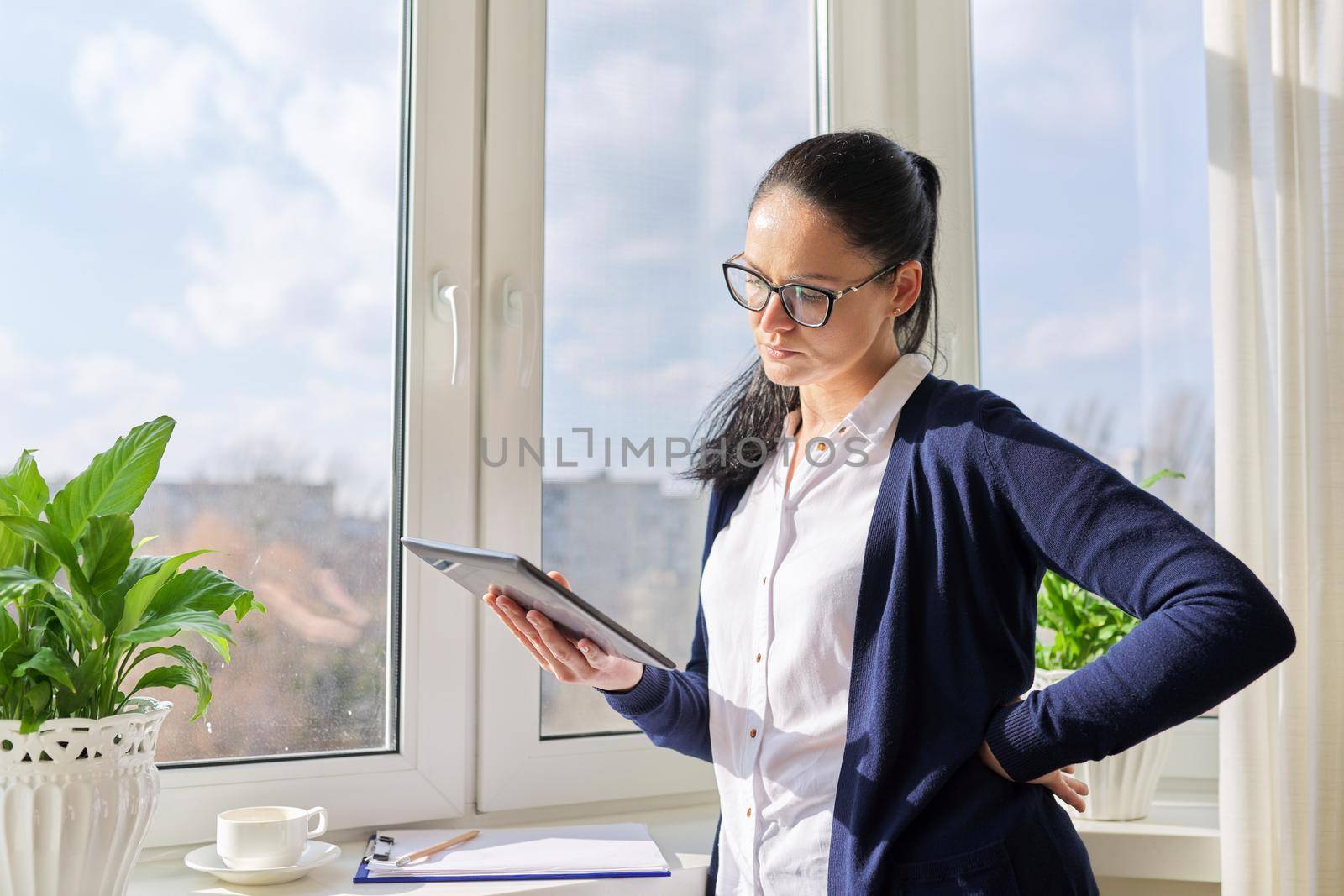Serious business woman working remotely in her home office, talking on video call, using digital tablet. Freelance, technology, telecommunications, business remotely, video communication near window
