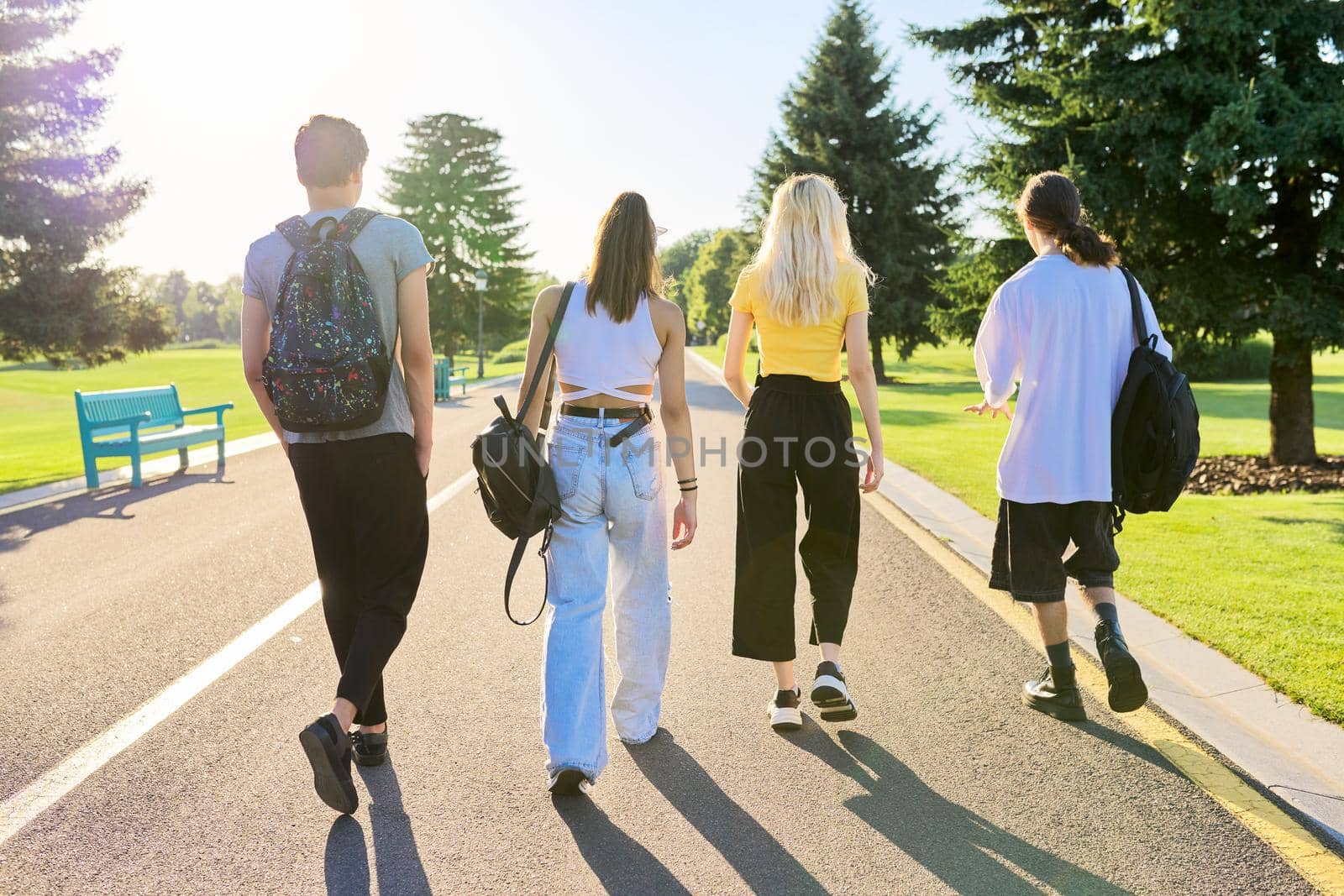 Outdoor, four teenagers walking together on road. Group of happy teenage friends on sunny summer day, back view. Adolescence, youth, friendship, young people, high school college concept