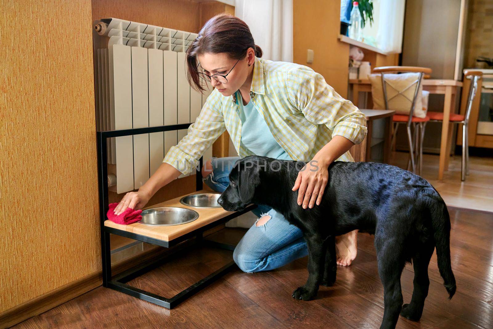 Middle aged woman and pet dog at home in kitchen interior. Pet owner cleaning near food bowls, black labrador puppy 4 months old. Lifestyle, love, pets, 40s people concept