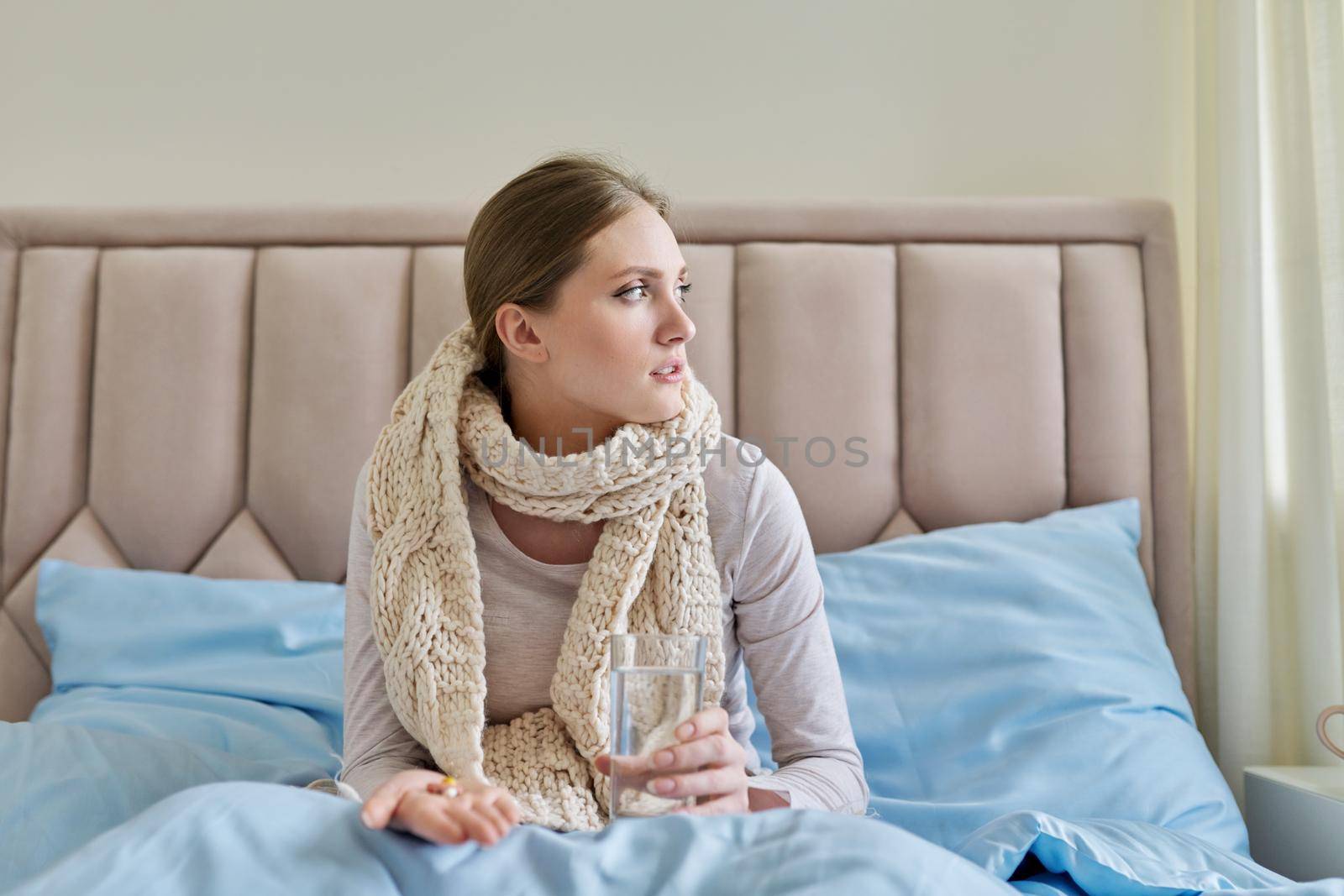 Sick woman with glass of water and pills in her hand, young female sitting at home in bed taking medicine. Medicine, pharmacology, flu season, seasonal diseases, people concept