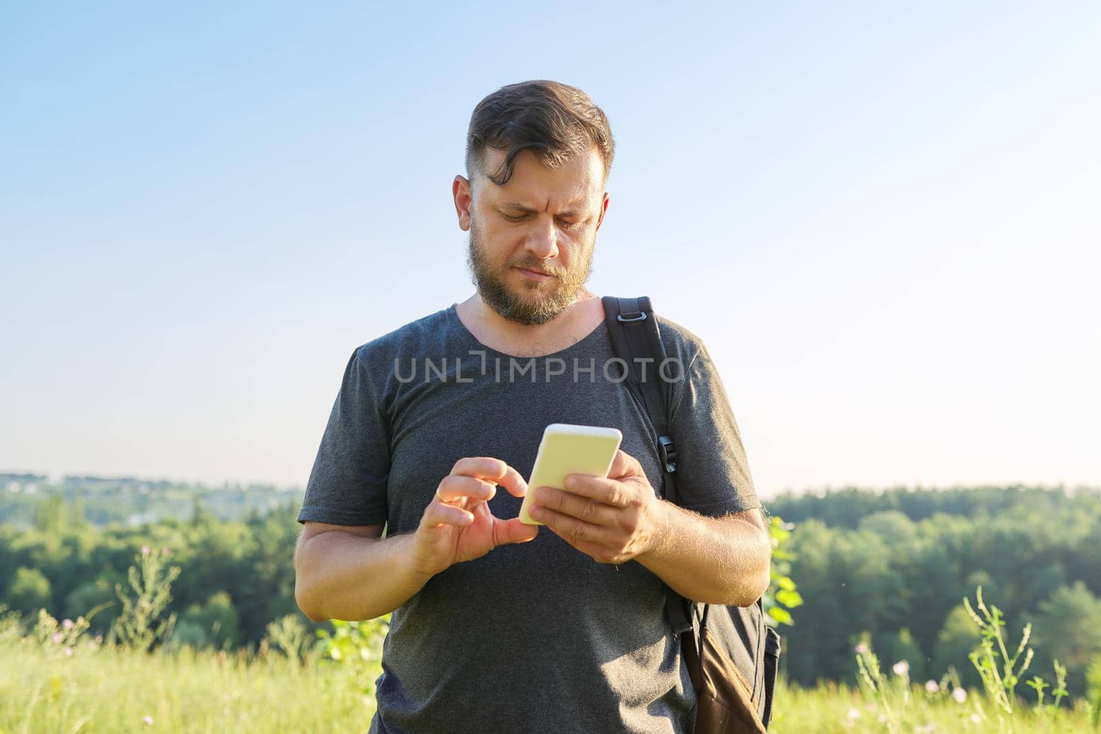 Middle-aged bearded man with backpack using smartphone. Summer sunny day, nature, sky background. Hike, adventure, vacation, technology, communication, leisure people concept