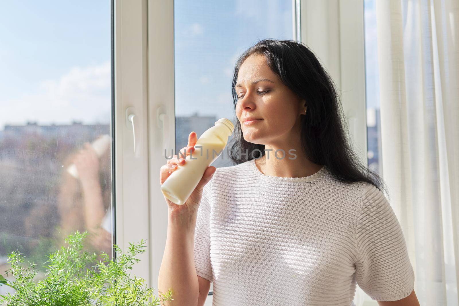 Woman drinking milk drink from bottle standing near window, milk yogurt dairy healthy drinks, copy space. Healthy food, diet, natural products