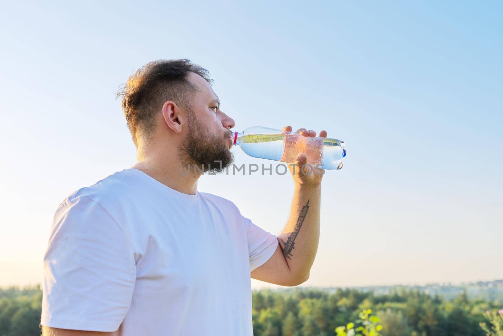 Middle-aged bearded man drinks water from bottle on hot summer day, nature meadow sky hills background. Summer, heat, health concept, copy space