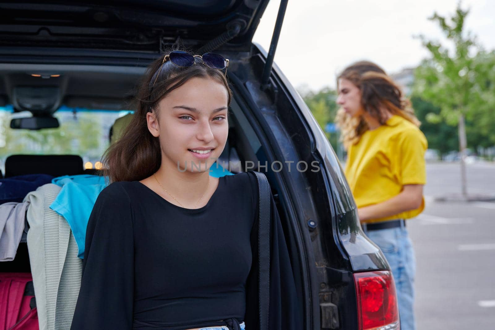 Portrait of a beautiful teenage girl of fourteen years. Adolescent female near the car, city guy friend background.
