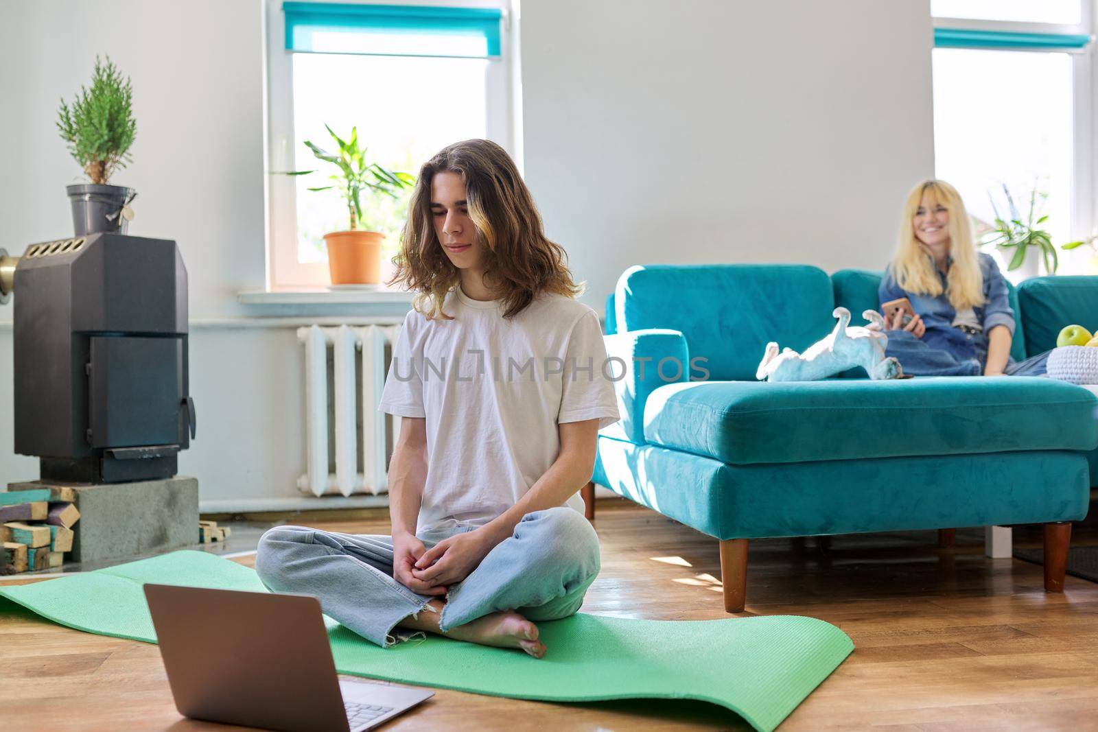 Guy teenager sitting in lotus position on yoga mat at home on floor with laptop. Meditation, online training, leisure, lifestyle, teenagers concept