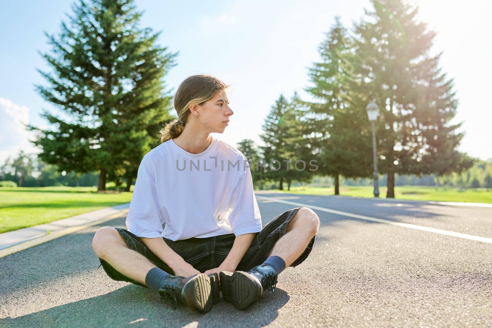 Portrait of handsome teenage guy, profile view outdoors, copy space. Serious hipster teenager with long hair wearing white t-shirt on road on sunny summer day. Youth, adolescence, people, lifestyle concept