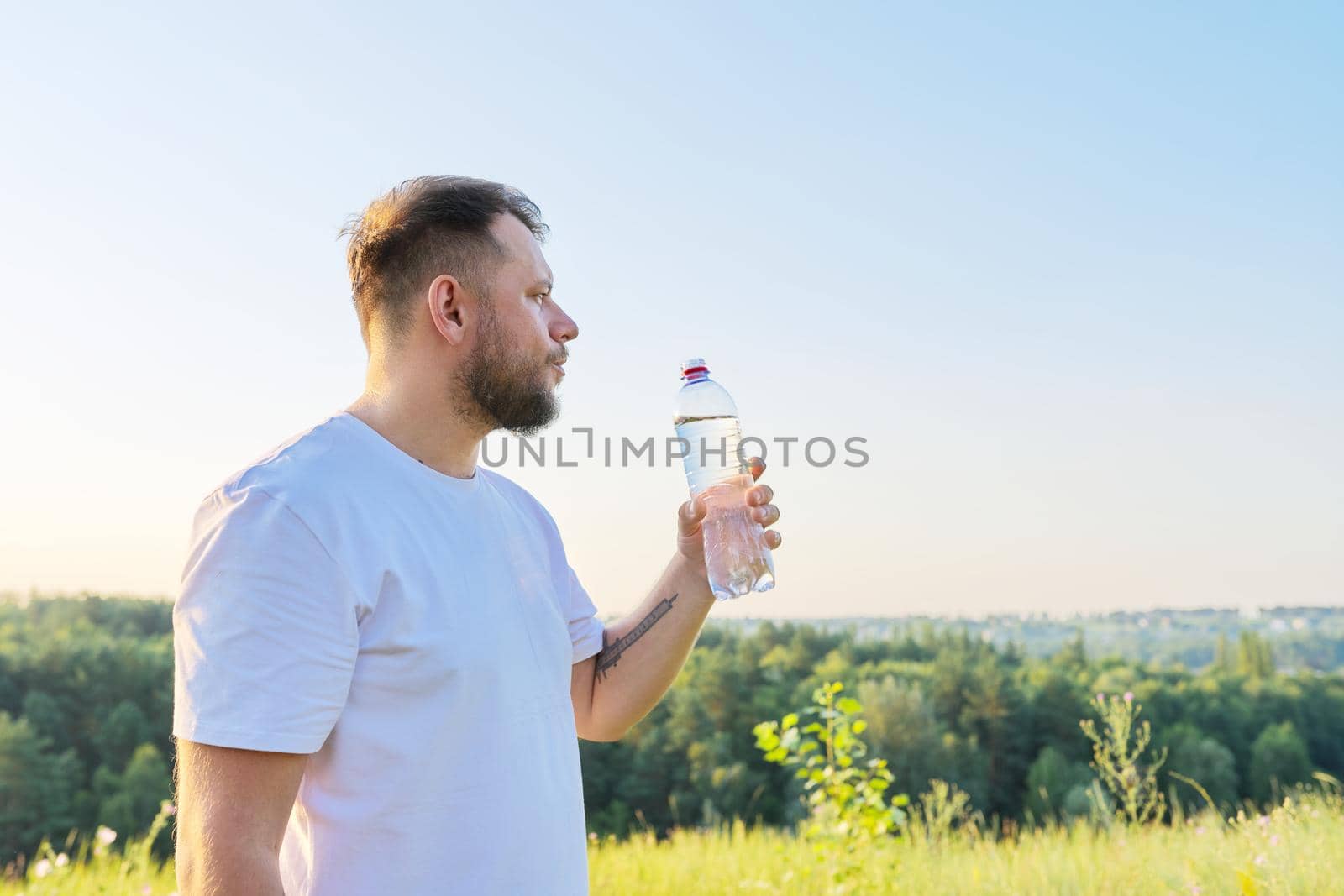 Middle-aged bearded man drinks water from bottle on hot summer day, nature meadow sky hills background. Summer, heat, health concept, copy space