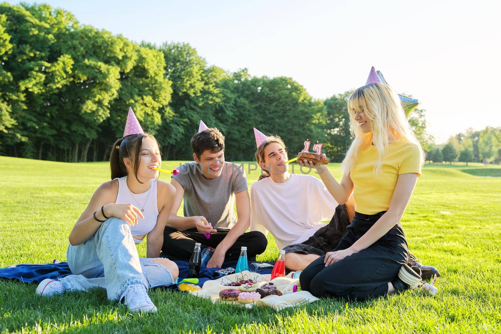 Birthday party. Teenager girl with cake with candles 17, celebrating birthday with friends, teenagers in festive hats sitting on grass in park, summer sunny day
