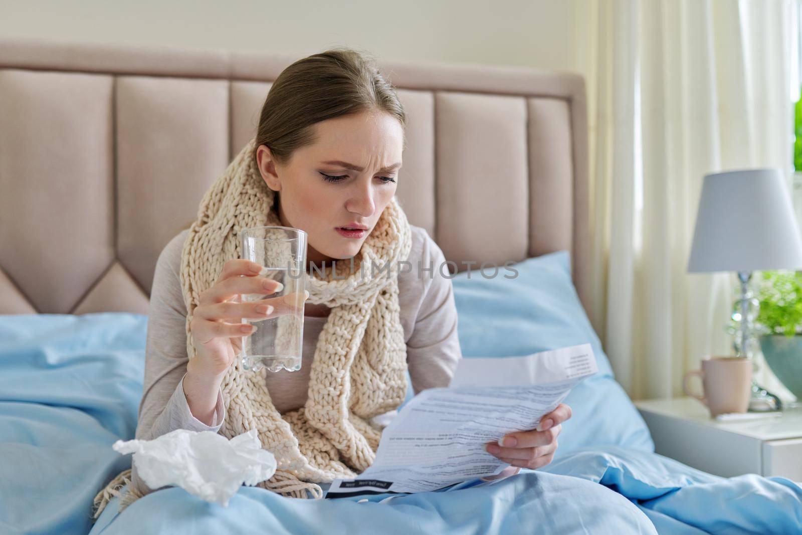 Young sick woman at home in bed, female with glass of water reading instructions and recommendations for medication. Season of colds, viral diseases, health, medicine, pharmacology, people concept.