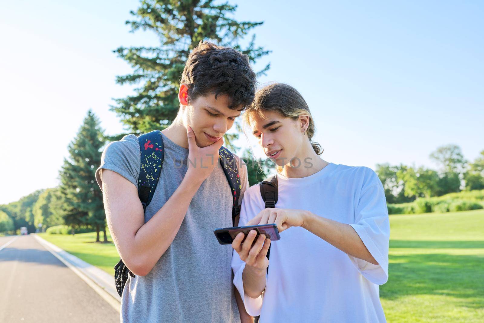 Teenagers friends talking looking at smartphone screen. Two guys 16, 17 years old with smartphone outdoors, on road on sunny summer day. Technology, lifestyle, friendship, youth, adolescence concept