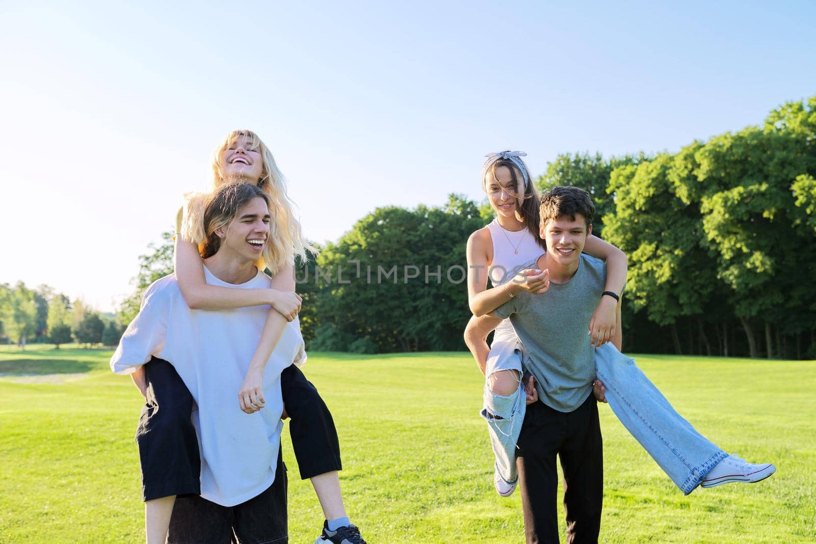 Happy group of teenagers having fun outdoors. Two young laughing couples of teenage friends, outdoors in park, green lawn grass background, sunny summer day. Adolescence, youth, friendship, young people